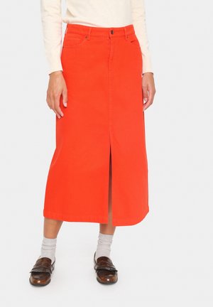Person iført et klart orange midi-nederdel med frontslids, hvid langærmet top, grå sokker og brune loafers, der står mod en ensfarvet baggrund.