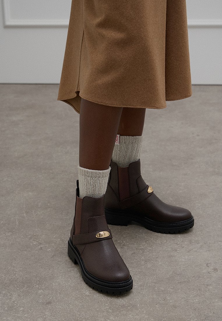 Botas de tobillo de cuero marrón con un acabado texturizado, acentuadas por una hebilla dorada y suelas de goma negra. Llevadas con calcetines color crema.