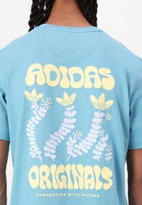 Zila kokvilnas T-kreklu ar lieliem dzelteniem uzrakstiem "ADIDAS" un "ORIGINALS", ko akcentē baltiem un dzelteniem ziedu grafiskajiem attēliem, veicinot dabas tēmu.