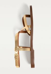 Guldmetallic strappy sandal med en brun flad sål. Har to krydsede remme og en sidebukke til justerbar pasform. Glat tekstur.