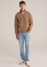 Felpa marrone con zip, tessuto leggero, maniche a coste, abbinata a jeans azzurri chiari e sneakers bianche con suola beige. Design semplice.