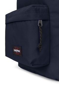 Námořnický modrý batoh vyrobený z odolné látky s přední kapsou, kovovým zipem a nášivkou loga Eastpak v červené a černé barvě. Texturovaný povrch.