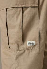 Beige cargo lomme med et rutenetmønster, syede sømme og et mærke med teksten "STANDARD ISSUE." Stoffet ser tekstureret og holdbart ud.