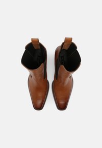 Bottines en cuir brun avec des panneaux latéraux en élastique, bout pointu et accents sombres texturés contrastants sur le dessus. Semelle plate.