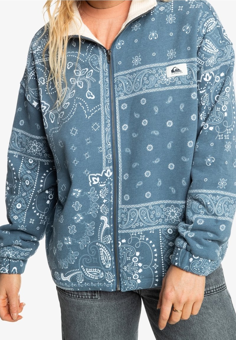 Mujer que lleva puesta una chaqueta azul con cremallera y un patrón de paisley blanco, además de un pequeño parche con logo en la parte superior del pecho, combinada con jeans grises.
