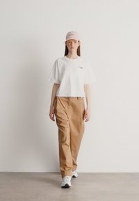 Camiseta blanca recortada con un pequeño logo, pantalones anchos color beige y una gorra rosa claro. Zapatillas blancas completan el conjunto. Fondo sencillo.