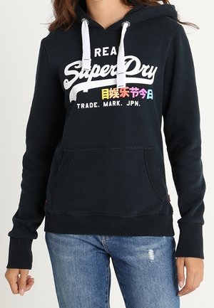 Osoba nosząca czarną bluzę z kapturem SuperDry z białymi sznurkami i kolorowym japońskim napisem nad "TRADE. MARK. JPN.", w połączeniu z niebieskimi dżinsami.