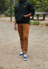 Felpa verde scuro con tasca frontale, pantaloni tapered color sabbia e sneakers multicolori con dettagli blu, rossi e bianchi.