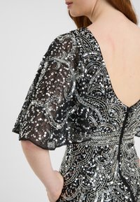 Abito nero con maniche trasparenti e leggere decorate con paillettes argentate e intricati ricami a perline in un motivo decorativo. Dettaglio della schiena bassa con chiusura a zip.