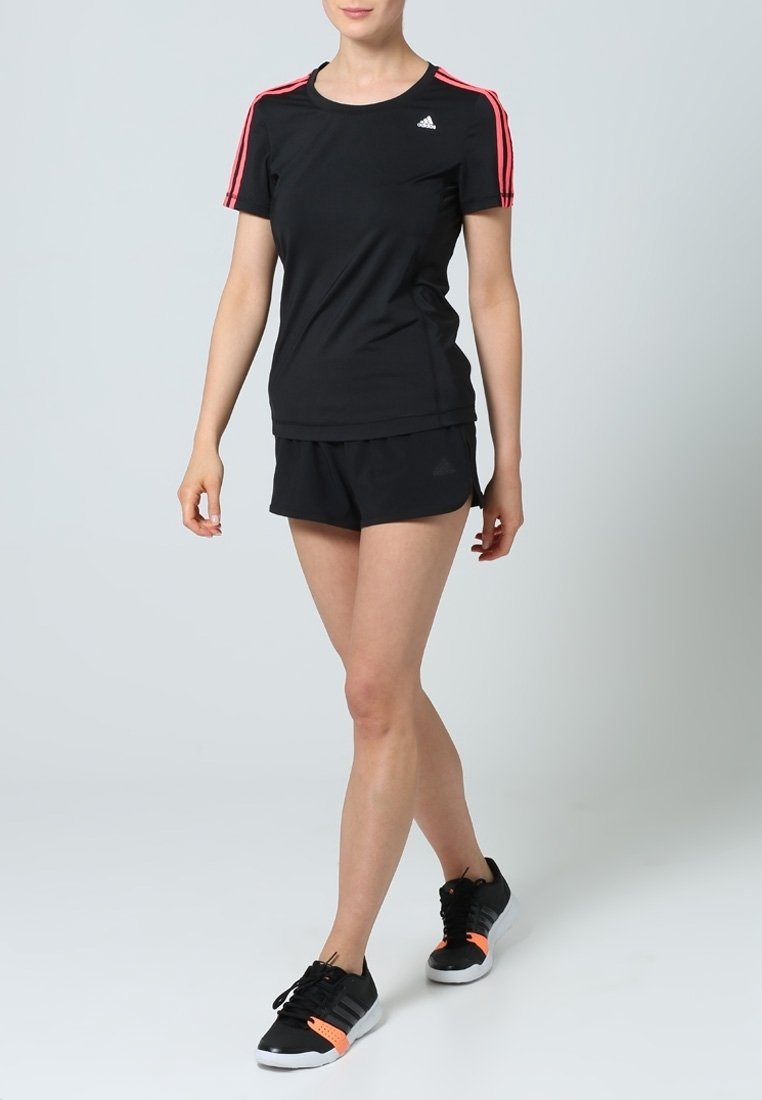 Femme portant une tenue de sport Adidas noire avec des bandes rouges sur les manches et des baskets noires, debout devant un fond clair uni.