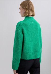 Grøn strikket sweater med høj hals, ribbet kant og tekstureret stof; har sænket skuldre og et stilfuldt bagdesign.