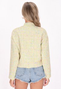Gevoerde gele jas met meerkleurige spikkels, met een cropped ontwerp en lange mouwen. Draag met denimshorts, weergegeven vanaf de achterkant.