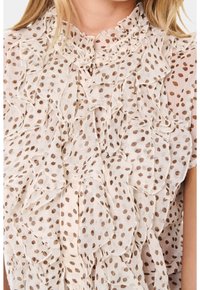 Camicia in crema trasparente con tessuto leggero, strati arricciati e pois marroni; include chiusura con bottoni al collo.