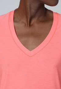 Persona de piel oscura que lleva una camiseta de canalé color coral claro con cuello en V, mostrando el cuello y la parte superior del pecho, mirando hacia la derecha.