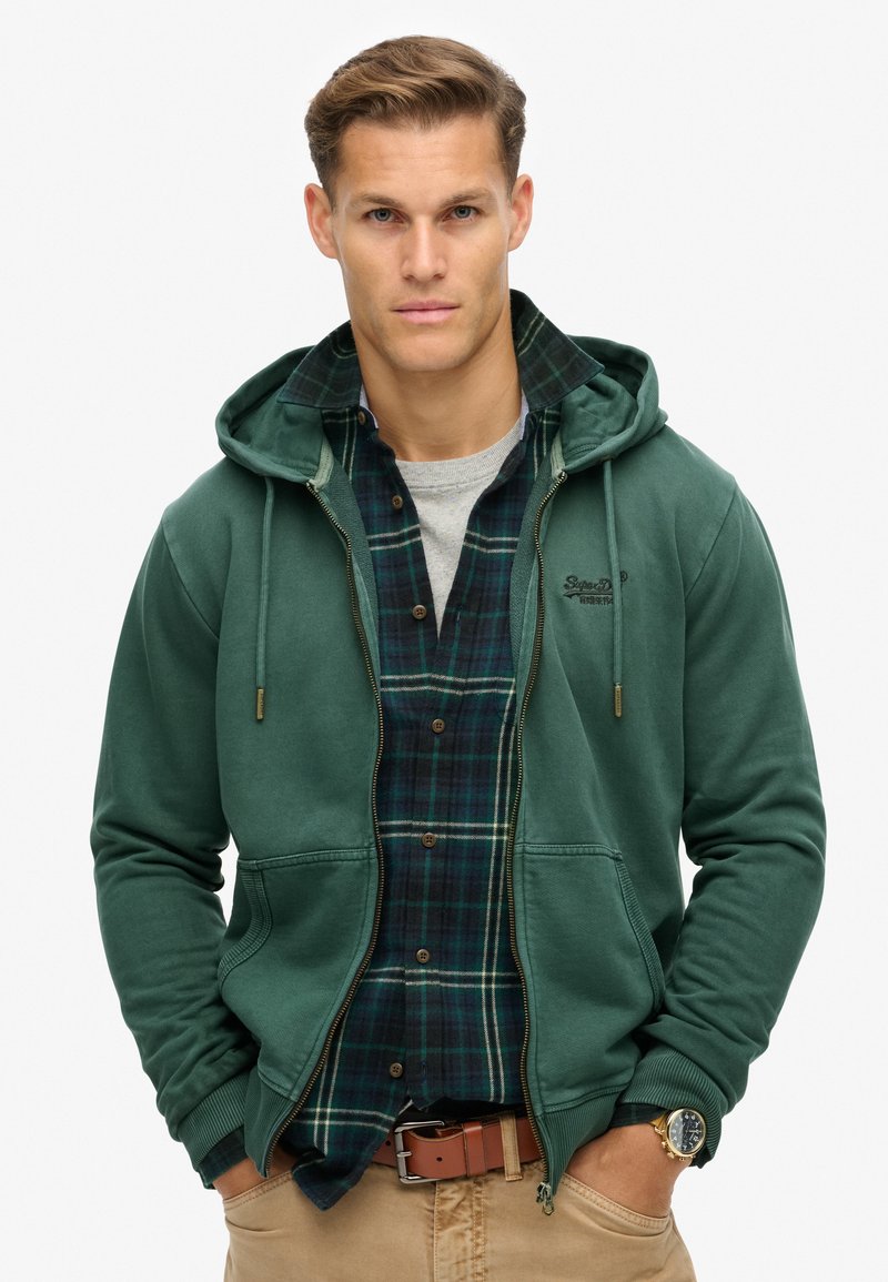 Sudadera verde con cremallera y acabado texturizado, llevada sobre una camisa de cuadros azul y verde. Acentuada con un logotipo, con capucha y bolsillos frontales.