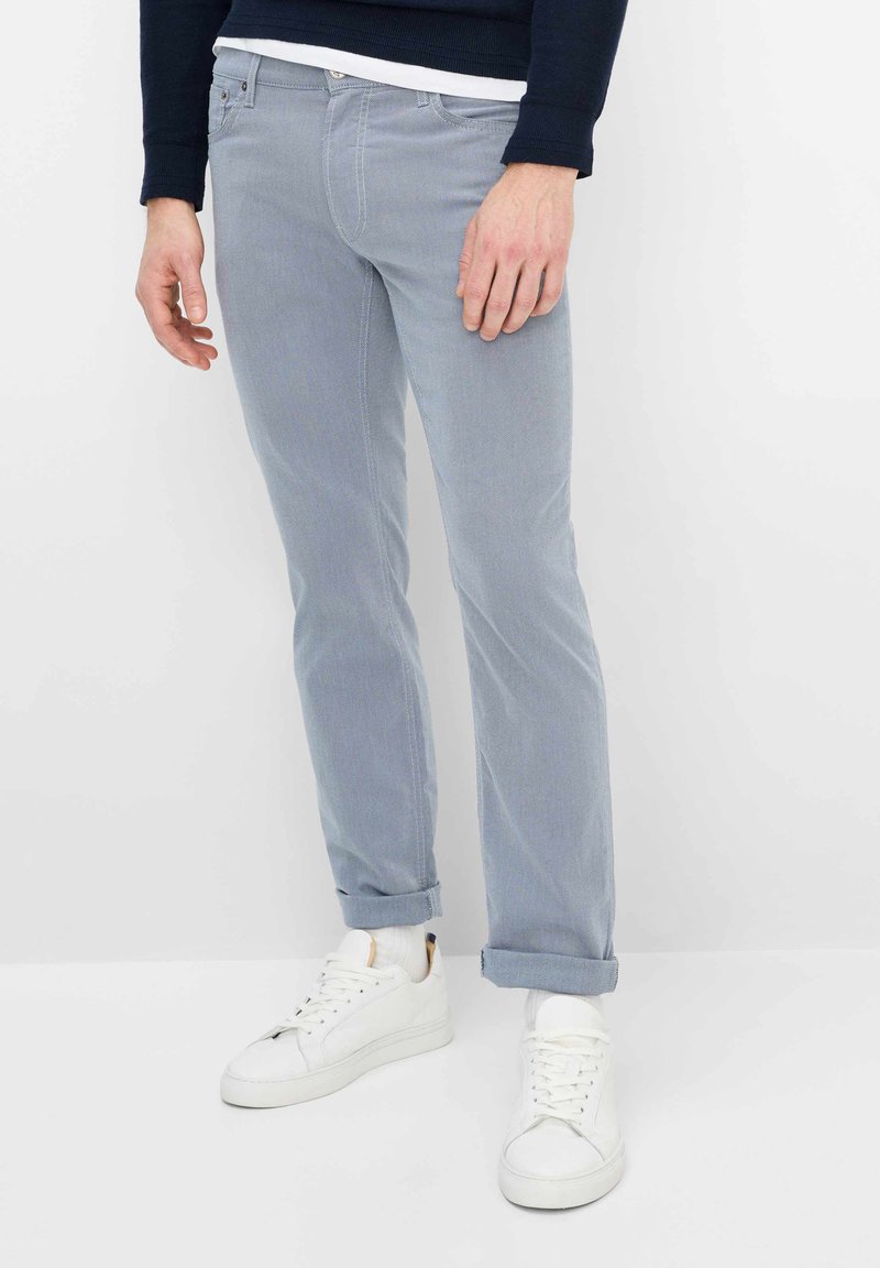 Man draagt lichtblauwe opgerolde broek, witte sneakers, witte sokken en een donker shirt met lange mouwen, staand tegen een witte achtergrond.
