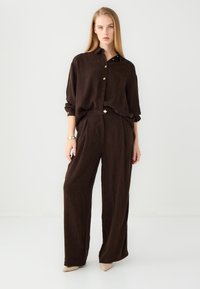 Braune, strukturierte Bluse mit goldenen Knöpfen, kombiniert mit weit geschnittenen braunen Hosen und beigen Absätzen; minimalistisches Design mit subtilen Details.