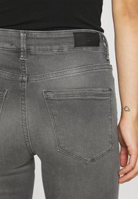Jeans cinzentos com um design ajustado, textura suave, cinco bolsos e um rótulo de cintura em couro preto. Costuras visíveis em linha contrastante.
