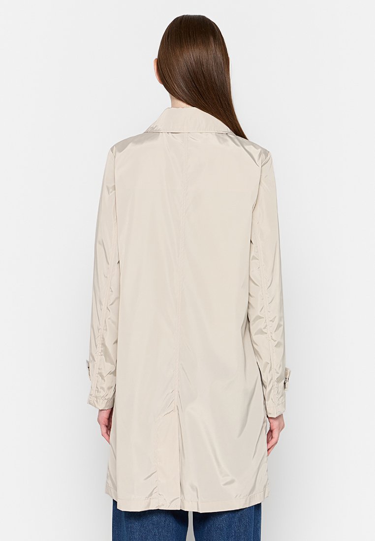 Femme aux longs cheveux bruns portant un trench-coat léger beige et un jean bleu, vue de dos contre un fond blanc uni.