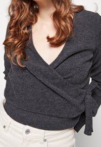 Czarny szary sweter o kroju kopertowym z głębokim dekoltem w literę V, teksturowana tkanina, długie rękawy i akcent w postaci wstążki w talii, zestawiony z białymi spodniami.