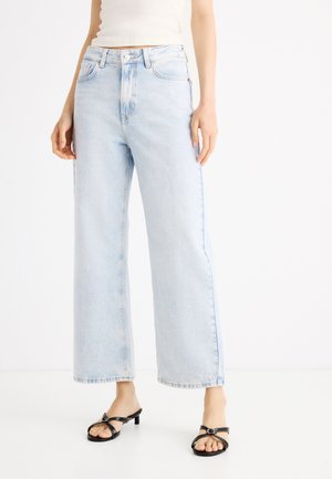 Kobieta ubrana w jasnoniebieskie szerokie dżinsy, białą bluzkę i czarne sandały na obcasie z paskami, stojąca na białej podłodze.