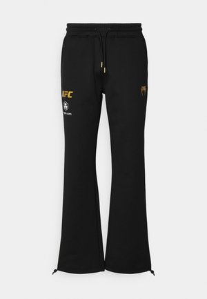 Svart joggebukse med en utsvingt design, laget av mykt stoff. har gulltrykt logo og justerbar snor i livet, med glidelåsdetaljer nederst.