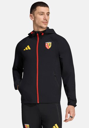 Uomo con giacca sportiva nera e pantaloni con cerniera rossa e loghi del club di calcio RC Lens, che guarda di lato con le mani in tasca.