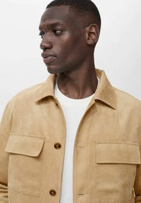 Beige suede jacket with a button front, two chest pockets, and a classic collar. Features a soft texture and a fitted silhouette.