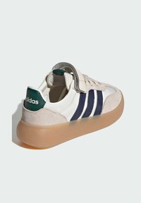 Zapatillas Adidas con una parte superior de cuero blanco, rayas en azul marino y acentos de gamuza beige. Suela de goma translúcida y marca verde en el talón.