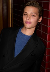 Schwarzer Nadelstreifenblazer über einem blauen Pullover, dazu ein schwarzes Einstecktuch. Die Person hat welliges, hellbraunes Haar und einen natürlichen Teint.