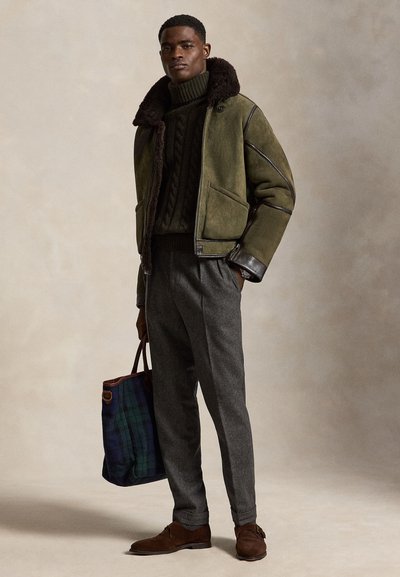 Mann trägt grüne Lammfelljacke, dunkelgrünen Zopfpullover, graue Hose, braune Schuhe, hält große karierte Tasche.
