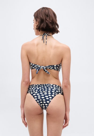 Γυναίκα με κοντά καστανά μαλλιά που φοράει μπλε ναυτικό μπικίνι με μεγάλα λευκά πουά, εμφανιζόμενη από πίσω σε απλό φόντο.