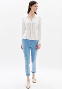 Witte blouse met lange mouwen en V-hals, versierd met knopen, gecombineerd met lichtblauwe slim-fit broek en gesloten hakken, beige sandalen met strepen.