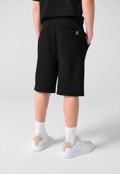 Personne portant un short noir mi-cuisses avec une petite étiquette arc-en-ciel sur la poche arrière, des chaussettes blanches et des baskets blanches, debout sur un sol uni.