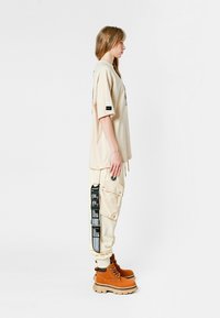 Camiseta beige de gran tamaño con mangas cortas, combinada con pantalones cargo beige claros que presentan detalles de parches negros y botas de encaje marrones.