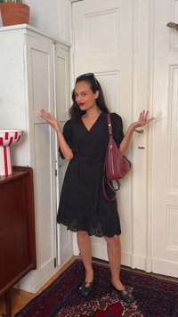 Γυναίκα σε μαύρο φόρεμα και μαύρα παπούτσια στέκεται με τα χέρια ψηλά, κρατώντας μια μπουρντό τσάντα σε ένα δωμάτιο με λευκές πόρτες και χαλί με σχέδια.