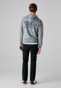 Felpa grigia con la scritta "RED BULL RACING" in stampa blu navy, progettata da Pepe Jeans. Indossata con jeans scuri e sneakers. Design semplice, vestibilità casual.