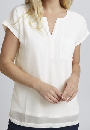 Femme portant un chemisier blanc à manches courtes avec une poche poitrine, une fente en V, et une bande horizontale transparente près de l'ourlet, les mains légèrement jointes.