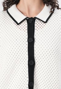 Bílé pletené kardigan s černým zapínáním na knoflíky. Obsahuje límec s černým lemem a strukturovaný, otevřený pletený design.