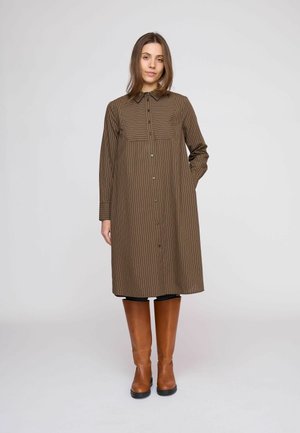 Donna che indossa un vestito midi marrone a righe con bottoni e stivali di pelle beige fino al ginocchio, in piedi davanti a uno sfondo grigio chiaro uniforme.