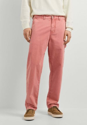 Persona que lleva vaqueros rosas desteñidos de corte holgado con bolsillo lateral y zapatos casuales marrones, de pie frente a un fondo claro y liso.