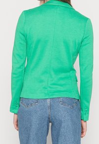 Blazer vert cintré en tissu texturé, avec un col à revers cranté et des accents de quatre boutons sur les poignets. Vue de dos avec coutures ajustées.