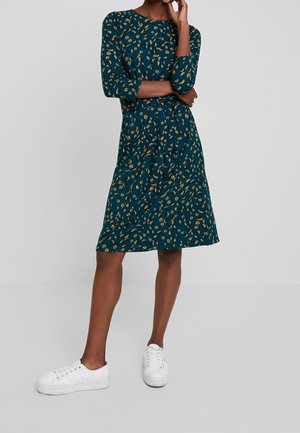 Persona con vestido verde azulado hasta la rodilla con patrón de hojas amarillas y mangas tres cuartos, combinado con zapatillas blancas, de pie con los brazos cruzados.