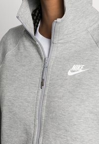Grå zip-hoodie i mjukt tyg med texturerad finish, med en vit Nike-logga på bröstet och hög krage.