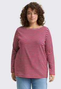 Vrouw met krullend haar, gekleed in een rood-witte horizontaal gestreepte longsleeve en lichtblauwe jeans, die recht vooruit kijkt tegen een effen achtergrond.