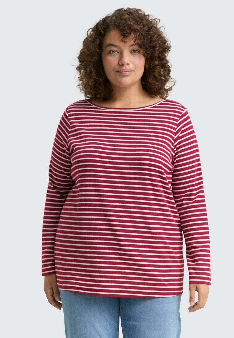 Vrouw met krullend haar, gekleed in een rood-witte horizontaal gestreepte longsleeve en lichtblauwe jeans, die recht vooruit kijkt tegen een effen achtergrond.