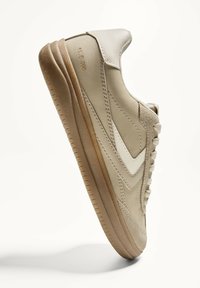 Beige sportssko med glat læder og ruskindsteksturer, med en lysfarvet sidestribe, snørebåndsdesign og gummisål.