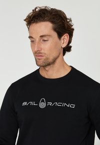 Schwarzer Langarm-Sweatshirt aus Baumwolle, mit gesticktem weißen "SAIL RACING"-Logo auf der Brust und Rundhalsausschnitt.
