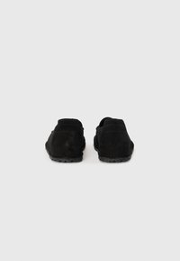 Sorte mockaskinnsko med lav profil, teksturert overdel, elastisk innsats og gummi såle med mønstrede grep. Baksidevisning som viser buet design.
