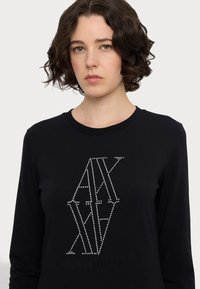 T-shirt nera a maniche lunghe in tessuto morbido, con un design geometrico realizzato in borchie argentate e la scritta "Armani Exchange" sottostante.
